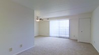 This single-story space features a spacious, open living area designed with simplicity and functionality in mind. The room is finished with wall-to-wall plush carpeting in a neutral beige tone, providing a soft and comfortable underfoot feel while contributing to the overall clean, understated aesthetic.

A prominent sliding glass door dominates one wall, dressed with full-length vertical blinds that allow flexible light control. This feature serves as the primary source of natural light, brightening the interior and offering a visual connection to the outdoors. When the blinds are drawn open, the room is bathed in daylight, enhancing the sense of openness.

The ceiling is smooth and finished in white, maintaining a bright, airy atmosphere throughout. A ceiling fan with an integrated light fixture is centrally positioned overhead, providing both ventilation and ambient lighting — a practical and permanent fixture suited to year-round use.

The walls are painted in a soft, neutral tone that complements the carpeting, creating a cohesive and versatile interior palette. White baseboards frame the perimeter of the room cleanly. The overall layout is open and unobstructed, allowing flexible furniture arrangement and making the space feel generously proportioned.