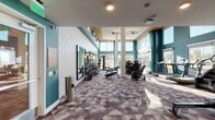 This single-floor fitness center is designed with both functionality and aesthetics in mind. The space is immediately striking thanks to its soaring ceilings, bold teal accent walls, and floor-to-ceiling windows that flood the interior with abundant natural light and provide open views of the surrounding landscape. Decorative white scalloped wall panels add a textural contrast, while a geometric-patterned carpet in muted tones of gray and mauve grounds the space with a modern sensibility. Structural columns with warm wood ceiling accents above anchor the main workout area, adding architectural interest throughout.

The fitness center is comprehensively equipped for a full range of workouts. A dedicated strength training zone features multi-station cable machines, adjustable benches, and a well-stocked dumbbell rack lined along mirrored walls. The cardio section includes treadmills, stair climbers, ellipticals, and rowing machines positioned near large windows to maximize natural light and outdoor views. Ceiling fans provide additional airflow throughout. A separate area offers additional functional fitness space, complete with a water station and built-in storage cubbies for convenience. Wall-mounted televisions are positioned throughout for entertainment during workouts. The overall ambiance is clean, energizing, and thoughtfully designed to support an active lifestyle.