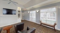 This single-floor communal space is thoughtfully designed to serve both relaxed gathering and focused meeting needs. The room is defined by vaulted ceilings with clean crown molding and soft gray walls that lend a calm, sophisticated atmosphere throughout. Large windows fitted with white plantation-style blinds line multiple walls, allowing generous amounts of natural light to fill the space while offering views of the surrounding greenery outdoors.

The lounge area features comfortable dark leather armchairs arranged around small round side tables, creating an inviting setting for informal conversation or quiet relaxation. A upholstered bench seat adds additional seating along the perimeter. A wall-mounted flat-screen television is positioned adjacent to a built-in shelving unit with illuminated display shelves and cabinet storage below, providing both functional and decorative appeal. Sconce lighting is placed throughout, supplementing the ambient light from recessed ceiling fixtures and a ceiling fan.

A dedicated dining or meeting nook is set apart within the space, accommodating a wood dining table surrounded by cushioned chairs and bathed in natural light from corner windows. A console table and a glass-paned interior door add further character. The rich, patterned brown carpet with an abstract wave motif anchors the entire space and ties together its warm, welcoming aesthetic.