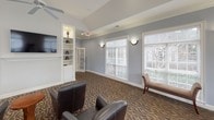 This single-floor communal space is thoughtfully designed to serve both relaxed gathering and focused meeting needs. The room is defined by vaulted ceilings with clean crown molding and soft gray walls that lend a calm, sophisticated atmosphere throughout. Large windows fitted with white plantation-style blinds line multiple walls, allowing generous amounts of natural light to fill the space while offering views of the surrounding greenery outdoors.

The lounge area features comfortable dark leather armchairs arranged around small round side tables, creating an inviting setting for informal conversation or quiet relaxation. A upholstered bench seat adds additional seating along the perimeter. A wall-mounted flat-screen television is positioned adjacent to a built-in shelving unit with illuminated display shelves and cabinet storage below, providing both functional and decorative appeal. Sconce lighting is placed throughout, supplementing the ambient light from recessed ceiling fixtures and a ceiling fan.

A dedicated dining or meeting nook is set apart within the space, accommodating a wood dining table surrounded by cushioned chairs and bathed in natural light from corner windows. A console table and a glass-paned interior door add further character. The rich, patterned brown carpet with an abstract wave motif anchors the entire space and ties together its warm, welcoming aesthetic.