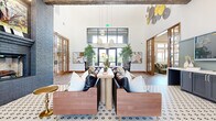Located at 1700 S Mayhill Rd in Denton, TX, this single-story community clubhouse offers a generously sized, thoughtfully designed amenity space. The grand entrance lounge immediately impresses with soaring ceilings, exposed wood beam accents, and abundant natural light streaming through large black-framed windows and glass doors. Stylish pendant lighting, curated wall art, and lush potted greenery create a welcoming, upscale atmosphere. Comfortable leather seating arrangements and patterned area rugs anchor the central lounge area, which also features an eye-catching decorative wall display with yellow-backed shelving and a painted brick fireplace surround. The fully equipped kitchen showcases sage-green cabinetry, gold fixtures, vertical subway tile, open wood shelving, and stainless steel appliances. Private conference rooms are finished with sophisticated dark grid-panel accent walls, round tables, and soft upholstered seating, while individual co-working nooks offer built-in desks with botanical wallpaper accents. The fitness center is bright and airy, lined with expansive windows, exposed beam ceilings, and a full suite of modern cardio equipment. Throughout, warm wood trim, herringbone carpet, and tile flooring unify the space with a refined yet approachable aesthetic.