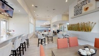 This single-floor community clubhouse offers a bright, open-concept layout designed for both relaxation and social interaction. The main lounge area is anchored by soaring ceilings and an elegant chandelier, with generous natural light streaming in through large windows. Comfortable seating arrangements—including plush sofas and accent chairs in neutral tones accented with pops of coral and orange—are arranged atop bold patterned area rugs, creating an inviting atmosphere for gathering.

A coastal-themed dining and café area features a striking large-format wall mural of an ocean pier, complemented by high-top tables and upholstered bar seating. The space transitions seamlessly into a well-appointed kitchen equipped with stainless steel appliances, white shaker cabinetry, quartz countertops, a kitchen island with bar seating, and mounted flat-screen televisions overhead.

Adjacent to the kitchen, a dedicated coffee and refreshment bar provides a self-serve station complete with a commercial coffee machine, microwave, refrigerator, and ample counter space, all finished with white cabinetry and light wood-look flooring consistent throughout the space.

The entry foyer is punctuated by a dramatic built-in planter filled with tall ornamental grasses, adding a natural, organic element to the clean, contemporary design. Light wood-look flooring, recessed lighting, and a cohesive coastal-modern aesthetic unite each area of this well-designed communal space.