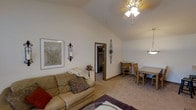 This single-story home showcases a comfortable and cohesive living space highlighted by a striking vaulted ceiling that adds a sense of openness and volume to the main living area. A ceiling fan with a light fixture is centrally mounted, providing both airflow and ambient lighting. The living room is finished with neutral beige carpeting that extends throughout, contributing to the warm and inviting atmosphere. Decorative wall accents, including an ornate iron sconce with a candle holder and a framed botanical artwork, add character to the space. A doorway leads to an adjacent room, suggesting a connected layout that enhances the flow of the home. Toward the right, a dining area is visible, furnished with a wooden table and matching chairs, illuminated by a pendant light fixture. The overall color palette of soft whites and warm beiges creates a cohesive, understated aesthetic throughout. The vaulted ceiling, decorative details, and open sightlines combine to give the space a welcoming and well-appointed feel suited for everyday living and casual entertaining.