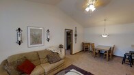 This single-story home showcases a comfortable and cohesive living space highlighted by a striking vaulted ceiling that adds a sense of openness and volume to the main living area. A ceiling fan with a light fixture is centrally mounted, providing both airflow and ambient lighting. The living room is finished with neutral beige carpeting that extends throughout, contributing to the warm and inviting atmosphere. Decorative wall accents, including an ornate iron sconce with a candle holder and a framed botanical artwork, add character to the space. A doorway leads to an adjacent room, suggesting a connected layout that enhances the flow of the home. Toward the right, a dining area is visible, furnished with a wooden table and matching chairs, illuminated by a pendant light fixture. The overall color palette of soft whites and warm beiges creates a cohesive, understated aesthetic throughout. The vaulted ceiling, decorative details, and open sightlines combine to give the space a welcoming and well-appointed feel suited for everyday living and casual entertaining.