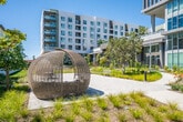 Located at 100 Zoo Ln in Santa Ana, California, this thoughtfully designed outdoor community space serves as a central gathering hub surrounded by contemporary multi-story residential buildings. The grounds are organized across a single level, offering a variety of distinct outdoor environments that cater to different activities and uses.

At the heart of the space lies a generous open lawn area framed by lush, professionally landscaped garden beds featuring ornamental grasses, flowering plants, shrubs, and mature trees. Wide, light-toned hardscape plazas provide ample room for social gatherings, complemented by seating benches and a distinctive woven gazebo structure that adds an artistic focal point to the courtyard.

Meandering concrete walkways lined with diverse plantings connect the various zones throughout the grounds, including a dedicated dog park enclosed by wrought iron fencing. Building entries are accessible via paved pathways flanked by vibrant garden plantings, with large floor-to-ceiling windows on the ground-floor facades allowing natural light to pour into interior spaces while maintaining a visual connection to the outdoors.

The overall ambiance is one of a well-maintained, park-like urban retreat designed to encourage community interaction. Under Southern California's abundant sunshine, the combination of greenery, hardscape, and modern architecture creates an inviting and functional outdoor environment.