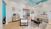 Located at 7466 Stephen Dr in Harrisburg, PA, this single-floor community facility offers a thoughtfully designed layout with a warm, welcoming ambiance throughout. A signature teal and soft blue color palette ties the space together, complemented by light wood-look flooring and an abundance of natural light streaming through multiple windows.

The combined dining, kitchen, and living area serves as the heart of the space. The dining area features a large dark wood communal table with high-back chairs and a wall-mounted TV, ideal for gatherings. Adjacent to it, the kitchen is outfitted with dark cabinetry, stone countertops, and modern appliances, accessible through a convenient pass-through window. The living room area is furnished with comfortable seating, decorative artwork, and elegant accents, creating a relaxed and inviting atmosphere.

The exercise room is generously sized and well-lit, housing multiple treadmills, a stationary bike, a rowing machine, and strength training equipment, all set on durable flooring with a wall-mounted TV for entertainment during workouts.

A private office nook provides a quiet workspace complete with a desk and computer setup. The facility also includes accessible restrooms. Throughout, the consistent use of teal accent walls, recessed lighting, and tasteful décor gives the space a cohesive, polished character.