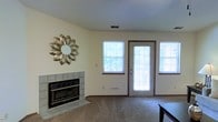 This single-story space centers around a comfortable living area designed with both functionality and warmth in mind. The room features a tile-surround fireplace as a focal point on one wall, providing a classic and practical heating element with clean, neutral finishes that complement the light-colored walls throughout. Adjacent to the fireplace, a decorative sunburst mirror adds a stylish accent to the space.

The living area is finished with wall-to-wall carpeting in a neutral tone, contributing to a warm and inviting atmosphere underfoot. Natural light flows generously into the room through multiple windows fitted with horizontal blinds, allowing for adjustable light control. A full-length glass exterior door further enhances the brightness of the space while providing direct access to the outdoors.

The layout is open and well-proportioned, offering a versatile arrangement for various furniture configurations. Ceiling-mounted HVAC vents indicate a central air system integrated throughout. The overall design is straightforward and practical, with neutral tones and traditional finishes that provide a clean, move-in-ready foundation suitable for a range of personal styles.