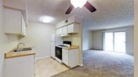 This single-floor residence features an open layout that seamlessly connects the kitchen and living area, creating a cohesive and functional living environment. The kitchen is equipped with white cabinetry, a gas range with oven, a refrigerator, and a built-in range hood. A butcher block-style countertop adds a warm, practical touch, while light vinyl flooring grounds the cooking space. Upper cabinets provide ample storage, and a ventilation unit is integrated into the ceiling above. The adjacent living area transitions to wall-to-wall carpeting, offering a comfortable and inviting atmosphere. A ceiling fan with a centered light fixture serves the living space, ensuring air circulation throughout. Textured ceilings are consistent across both areas, contributing to a cohesive aesthetic. A sliding glass door at the far end of the living room allows natural light to filter in while providing access to an outdoor area. The neutral color palette of soft grays and whites throughout keeps the space feeling clean and understated, making it well-suited for a variety of interior styles.