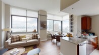 This thoughtfully designed high-rise residence offers sweeping panoramic views of the city skyline and waterfront through expansive floor-to-ceiling windows that flood the interior with natural light. The open-concept living and dining area forms the heart of the home, where large corner windows frame striking skyline vistas. The living room features warm hardwood flooring, neutral tones, and generous proportions, while the adjacent dining area accommodates a glass-top table positioned to take full advantage of the surrounding views. The kitchen is outfitted with rich dark wood cabinetry, granite countertops, and tile flooring, and includes direct access to a private outdoor balcony overlooking the urban landscape. The bedroom is a serene retreat with its own wall of floor-to-ceiling windows offering remarkable waterfront and skyline views, light hardwood floors, and a walk-in closet. Two bathrooms serve the residence: a full bathroom with a soaking tub, dark wood vanity, and clean white tile, and a half bathroom distinguished by deep purple accent walls, a dark wood vanity, and stylish framed artwork. Throughout the space, a consistent palette of warm neutrals and rich wood tones creates a cohesive and sophisticated ambiance.