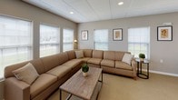 This single-floor communal facility is thoughtfully designed to support both relaxation and an active lifestyle. The space is organized into distinct functional areas, each finished with a consistent neutral palette of warm grays and beige tones that create a cohesive, welcoming atmosphere throughout.

The lounge area is a standout feature, centered around a large L-shaped sectional sofa in a warm tan upholstery that wraps generously around a rustic wood-and-metal coffee table. Multiple windows fitted with white horizontal blinds line the walls, allowing abundant natural light to fill the room. Framed botanical artwork accents the walls, adding a refined touch to the casual setting.

Adjacent to the lounge is a fully equipped kitchen, featuring stainless steel appliances including a refrigerator, electric range, microwave, and dishwasher. White cabinetry pairs cleanly with dark countertops, and warm hardwood-style flooring grounds the space. A peninsula counter with additional seating extends into a casual sitting area furnished with dark leather club chairs.

The fitness center is well-appointed with cardio equipment including treadmills, ellipticals, and a stationary bike, alongside a multi-function cable strength machine and an adjustable weight bench. A wall-mounted television and natural light from multiple windows make the space practical and inviting for regular use.