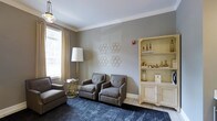 This single-floor space presents a thoughtfully designed sitting area that balances sophistication with comfort. The room is finished with crown molding along the ceiling line, adding a refined architectural detail that frames the space elegantly. Walls are painted in a soft, neutral gray tone, creating a calm and polished backdrop throughout.

Natural light enters through a window dressed with sheer and gold-toned drapery panels, lending warmth to the overall palette. The seating arrangement features a pair of tufted gray armchairs positioned symmetrically on a deep navy area rug, complemented by an additional upholstered chair and a gold-accented side table. A tall floor lamp provides ambient lighting alongside a vintage-style pendant fixture overhead.

A standout feature is the ornate cream-colored display cabinet with nail-head trim detailing, showcasing decorative gold vessels and curated accessories across its open shelving. Geometric hexagonal metal wall art adds a contemporary visual element against the neutral wall. The combination of classic furnishings, metallic accents, and layered textures creates a cohesive and inviting atmosphere well-suited for a communal or reception-style lounge area.