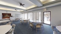 This single-floor clubhouse is a well-appointed community gathering space designed to accommodate a variety of events and functions. The main hall is generously sized, featuring a combination of blue carpet and large-format tile flooring that visually defines distinct areas within the room. Multiple round tables with folding chairs are arranged throughout, offering flexible seating configurations for group gatherings. The ceiling is a notable architectural feature, with a recessed tray design accented by ambient cove lighting and complemented by multiple ceiling fans to ensure comfortable airflow throughout. Large windows with vertical blinds line one wall, allowing natural light to fill the space and offering views to the exterior. A wall-mounted flat-screen television and a fireplace with a gold-trimmed surround serve as focal points along the brick-accented feature wall, adding warmth and character to the lounge area. Adjacent to the main hall is a fully equipped kitchen, outfitted with warm wood cabinetry, a mosaic tile backsplash, dark countertops, and stainless steel appliances including a full-size refrigerator, range, and microwave. The tile flooring in the kitchen provides a clean, practical surface. Together, these spaces create a functional and inviting environment suited for social events, meetings, and community activities.