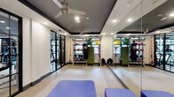 This fitness studio is thoughtfully designed to support a wide range of workout styles within a single, open-plan space. The room features a two-tone ceiling with recessed lighting and dual ceiling fans, ensuring the area remains well-lit and ventilated during intense training sessions. A full-length mirrored wall runs along one side of the room, visually expanding the space while allowing users to monitor their form during exercise.

The flooring consists of speckled terrazzo-style material, offering a durable and easy-to-clean surface suited for high-activity use. Equipment is well-organized throughout the space, including a multi-rack storage unit stocked with medicine balls, kettlebells, and resistance bands. A heavy punching bag, stability balls in multiple sizes, and a cable/functional training rig add versatility for strength, cardio, and functional fitness routines.

Padded blue exercise benches are positioned toward the front of the room, providing space for stretching or floor-based exercises. Large black-framed glass doors at the entrance allow natural light to filter in, creating an inviting and energizing atmosphere. Overall, the studio balances functionality with a clean, modern aesthetic.