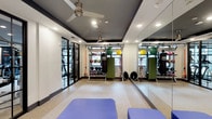 This fitness studio is thoughtfully designed to support a wide range of workout styles within a single, open-plan space. The room features a two-tone ceiling with recessed lighting and dual ceiling fans, ensuring the area remains well-lit and ventilated during intense training sessions. A full-length mirrored wall runs along one side of the room, visually expanding the space while allowing users to monitor their form during exercise.

The flooring consists of speckled terrazzo-style material, offering a durable and easy-to-clean surface suited for high-activity use. Equipment is well-organized throughout the space, including a multi-rack storage unit stocked with medicine balls, kettlebells, and resistance bands. A heavy punching bag, stability balls in multiple sizes, and a cable/functional training rig add versatility for strength, cardio, and functional fitness routines.

Padded blue exercise benches are positioned toward the front of the room, providing space for stretching or floor-based exercises. Large black-framed glass doors at the entrance allow natural light to filter in, creating an inviting and energizing atmosphere. Overall, the studio balances functionality with a clean, modern aesthetic.