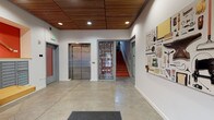 This lobby serves as the welcoming entry point of a modern, thoughtfully designed building. The space immediately impresses with its warm slatted wood ceiling, which contrasts elegantly against clean white walls and polished concrete floors, creating a contemporary yet inviting aesthetic. Recessed lighting illuminates the area evenly, enhancing the open, airy feel.

The entry side features wood-framed glass doors that allow natural light to filter in, offering views of the surrounding streetscape. A built-in wooden bench-style ledge runs along one wall, providing a functional yet stylish spot for packages or seating. Nearby, a bank of individual mailboxes is neatly integrated into the wall, accompanied by a vibrant orange accent wall displaying a decorative hammer installation.

A stainless steel elevator and stairwell provide vertical access throughout the building. A striking large-format typographic mural referencing the building's identity as "Kiln," a passive house development on North Williams, serves as a bold focal point. Additional wall-mounted artwork depicting an assortment of tools reinforces the building's craft-inspired character. Overall, the lobby strikes a balance between functionality and artistic expression, reflecting the building's energy-efficient, community-oriented design philosophy.