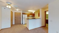 This single-floor space showcases an open-concept layout that seamlessly connects the kitchen and living area. The kitchen is equipped with medium-toned wood cabinetry offering ample storage, a white refrigerator with an ice and water dispenser, and a built-in electric cooktop with an overhead range hood. A peninsula-style countertop with a dark surface extends into the living space, creating a natural division between the two areas while maintaining an open feel.

A pendant light hangs above the peninsula, complementing the flush-mount ceiling fixture and a ceiling fan that serve the adjacent living area. The warm yellow-toned walls pair with light beige carpeting in the living space, transitioning to hard flooring in the kitchen zone. White baseboards and trim provide clean contrast throughout.

The entry door, finished in medium wood tones, is positioned directly across from the kitchen, offering a straightforward and functional layout. A hallway visible to the right suggests access to additional rooms. The overall ambiance is practical and welcoming, with a cohesive warm neutral palette throughout.