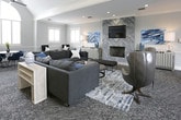 This refined single-floor lounge space makes an immediate impression with its soaring vaulted ceilings and a dramatic arched fanlight window that floods the interior with abundant natural light. The soft grey palette, wainscoting, and crown molding throughout lend a polished, classic aesthetic that is both welcoming and sophisticated.

The main lounge area is generously proportioned, anchored by a statement fireplace clad in textured chevron-patterned stone tile, above which a mounted television is framed by vibrant blue abstract artwork. A large sectional sofa in charcoal, a tufted leather bench, and a round coffee table define a comfortable central gathering zone, complemented by a patterned area rug and ceiling fans that enhance the airy feel.

Adjacent seating vignettes offer flexible arrangements, including a round dining table with contemporary blue chairs positioned beneath the arched window, and a pair of wingback armchairs suited for quieter conversation. A dedicated workspace or meeting area features a sleek rectangular desk with additional seating, set against a railing overlooking the lower level.

Plantation shutters on every window provide light control while maintaining the space's bright, open character. Recessed lighting supplements the natural illumination, ensuring the space remains inviting at all hours. The overall ambiance is one of curated comfort and understated elegance.