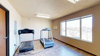 This single-floor fitness space offers a straightforward and functional environment designed for exercise and physical activity. The room is equipped with two treadmills positioned to take full advantage of the space, providing ample room for users to work out comfortably. The flooring consists of durable tile in a neutral, earthy tone that is both practical and easy to maintain. A large window along one wall floods the room with natural light and frames a view of the surrounding outdoor area, creating an open and airy atmosphere despite the room's modest footprint. The walls are finished in a soft, neutral beige tone, contributing to a calm and focused workout environment. Overhead, a flush-mounted fluorescent light fixture supplements natural lighting, ensuring the space is well-lit at any time of day. The textured ceiling adds a subtle architectural detail consistent with the room's clean, utilitarian aesthetic. A wall-mounted unit is also present, likely serving as a thermostat or control panel, supporting comfort during exercise. Overall, this fitness room is a practical and well-appointed space suited for light to moderate cardio workouts.