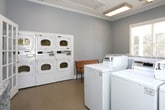 This shared laundry facility is a practical, well-organized space designed to accommodate multiple users efficiently. The room is finished with soft gray walls, crisp white trim, and crown molding, giving it a clean and polished appearance. Light tile flooring runs throughout, making the space easy to maintain.

The laundry area is outfitted with multiple top-load washing machines and a bank of stacked front-load dryers, providing ample capacity for high-volume use. A granite-topped folding counter runs along one wall, offering a generous surface for sorting and folding laundry. A utility sink is also present, adding further functionality.

Natural light filters in through several windows fitted with white horizontal blinds, keeping the space bright and airy. Overhead, recessed fluorescent light fixtures with warm wood-trimmed frames provide additional illumination. A wooden slatted bench with wrought iron legs offers a comfortable place to wait, and a waste receptacle is conveniently positioned nearby.

The room is accessible through a glass-paned French door that connects to an adjacent corridor. Overall, the space is thoughtfully arranged to balance utility and comfort, making it a well-appointed communal laundry facility.