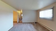 This single-floor, two-bedroom, one-bathroom unit offers a straightforward and functional layout designed for comfortable everyday living. The main living area is finished with wall-to-wall carpeting in a neutral tone, providing a soft and cohesive base throughout the space. Walls are painted in a clean, off-white palette that keeps the interior feeling bright and open.

A window along the exterior wall allows natural light to filter into the living room, illuminating the space during daytime hours. Baseboard heating runs along the perimeter, offering an efficient and consistent heat source throughout the colder months. The ceiling is smooth and white, complementing the overall clean aesthetic of the unit.

A hallway connects the living area to the rest of the unit, leading toward the two bedrooms and the shared full bathroom. Warm wood-toned door trim adds a subtle traditional detail to the otherwise minimalist interior. The layout flows logically from the main living space into the private areas of the unit, making efficient use of the available footprint. Overall, the space presents a clean, move-in-ready environment suited for a range of occupants.