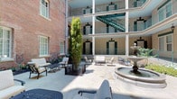 The Wash House Courtyard is a distinctive open-air space that seamlessly blends historic architectural character with modern amenity. The courtyard is enclosed by a multi-story building facade featuring wraparound balconies with decorative green shutters and iron railings, alongside an original exposed brick wall that lends the space a sense of historic authenticity. A central stone fountain serves as the focal point of the outdoor area, surrounded by a generously sized paved terrace finished in light-toned concrete. Seating is thoughtfully arranged throughout, with iron and upholstered lounge chairs and tables positioned to encourage gathering and relaxation. Tall, columnar evergreen shrubs and iron-fenced planter beds filled with lush tropical foliage introduce verdant contrast to the hardscape. An exterior staircase provides access to upper-level balconies, enhancing the courtyard's sense of vertical scale. Transitioning indoors, the ground-floor corridor features beige paneled walls accented with green trim details, pendant and wall-mounted lantern lighting, and a stainless steel elevator. Additional planter beds with broad-leafed greenery line the interior walkway, maintaining the botanical character established in the courtyard and creating a cohesive, inviting atmosphere throughout.
