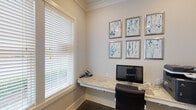 This single-level business center is designed to provide a functional and polished workspace. The centerpiece of the room is a wraparound, wall-mounted desk with a granite or stone-patterned surface that extends along two walls, offering generous workspace for computers, printers, and office equipment. The floating desk design contributes to a clean, uncluttered aesthetic while maximizing floor space.

Natural light is a defining feature of this space, with multiple large windows fitted with white horizontal blinds that allow for adjustable light control. The windows offer views of greenery outside, creating a pleasant connection to the outdoors while working.

The walls are finished in a soft, neutral tone that enhances the bright and airy atmosphere of the room. Crown molding adds an architectural detail that elevates the overall refinement of the space. A collection of six silver-framed botanical prints in soft blue tones is displayed on the wall above the desk, adding a decorative and cohesive visual element to the workspace.

The flooring is a neutral-toned carpet that adds warmth underfoot. A paneled white door provides privacy and separation from adjacent areas. The overall ambiance is calm, professional, and well-appointed, making it a practical yet aesthetically considered working environment.