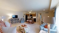 This single-floor residence offers a thoughtfully designed open-concept layout that seamlessly connects the living area and kitchen, creating a spacious and inviting atmosphere. The living area is generously sized, finished with plush carpeting that adds warmth underfoot, and furnished with comfortable seating arrangements centered around a flat-screen television housed on a classic cross-hatched media console. Natural and recessed lighting illuminate the space throughout, contributing to a bright and airy feel. The kitchen is a standout feature, showcasing rich wooden cabinetry, dark granite countertops, and a central island with bar seating — ideal for casual dining or entertaining. Black appliances, including a refrigerator, oven, and built-in microwave, provide a sleek contrast to the warm tones of the cabinetry. A tasteful tile backsplash adds a refined finishing touch to the culinary space. A hallway visible from the main living area suggests additional rooms beyond, hinting at a well-organized and functional floor plan. Overall, the residence balances comfort and practicality, offering a cohesive and livable environment with a clean, neutral palette that is easy to personalize.