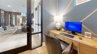 The Keegan Work From Home space is a thoughtfully designed private office environment that blends modern aesthetics with practical functionality. The focal point of the room is a striking blue-grey accent wall adorned with bold geometric gold-stripe detailing, creating a visually dynamic backdrop that sets the space apart. Light wood-tone flooring runs throughout, complementing the warm natural wood furnishings, including a generously sized desk with drawer storage and a coordinating side dresser. A stylish table lamp with a sculptural white base provides ambient task lighting, while a small window allows a touch of natural light to filter in. A built-in shelving unit with dark brick-tile backing offers open display and storage space, accessorized with books, decorative objects, and greenery. A striped upholstered bench beneath the shelving adds a casual seating option. A large chalkboard panel serves as both a functional message board and a design element. The space is enclosed by a full-length glass-panel door with a sleek black metal frame, offering privacy while maintaining visual connection to an adjacent lounge area visible beyond. The overall ambiance is modern, curated, and conducive to focused, comfortable work.