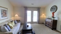 This single-floor residence offers a comfortable and well-appointed living environment designed with a clean, neutral aesthetic. The living room serves as the centerpiece of the space, featuring plush wall-to-wall carpeting that adds warmth underfoot. A standout feature is the set of French doors that flood the room with abundant natural light while providing access to an outdoor area, framed by elegant floor-length curtains in a soft, muted tone. The walls are finished in a warm off-white palette, creating a bright and airy atmosphere throughout. The living area is thoughtfully arranged with ample space for seating and a central coffee table, making it well-suited for both relaxing and entertaining. Flanking floor lamps provide supplemental warm lighting, enhancing the room's inviting ambiance. A wooden storage cabinet or credenza anchors one side of the room, adding a touch of functional elegance. The overall design is understated and versatile, offering a neutral canvas that accommodates a variety of furnishing styles. The single-level layout ensures seamless flow and accessibility throughout the home.