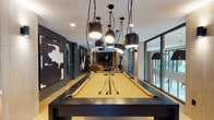 Located at 3560 Rambla Pl in Santa Clara, California, this single-floor amenity lounge is a well-appointed communal space designed with a modern, upscale aesthetic. The layout is elongated and open, seamlessly connecting distinct activity zones through light hardwood flooring and a cohesive dark-and-neutral color palette.

At the heart of the space sits a full-size billiards table in striking charcoal and gold tones, positioned beneath a dramatic cluster of large black pendant lights that serve as both a functional and sculptural focal point. Floor-to-ceiling glass windows line one side of the room, flooding the billiards area with abundant natural light and offering views of the surrounding outdoor environment.

Adjacent to the game area, a comfortable lounge zone features gold and mustard-toned sofas, mid-century-inspired accent chairs in warm caramel leather and cool gray upholstery, and a patterned area rug anchoring the seating arrangement. Tall indoor plants and bold abstract artwork mounted on deep slate-gray accent walls add visual depth and a curated, gallery-like character to the space.

A built-in media station with a wall-mounted flat-screen TV, a floating white counter, and backless wooden stools provides a casual viewing and workspace nook. Vertical wood slat paneling near the TV wall introduces warmth and texture. Recessed and wall-mounted lighting throughout ensures a well-lit yet intimate atmosphere suited for both social and leisure activities.