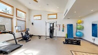 Located at 515 Aurora in Bend, Oregon, this dedicated exercise room offers a well-designed fitness environment thoughtfully laid out for a complete workout experience. The space features soaring ceilings with exposed wood beam accents, recessed lighting, and a row of wood-framed windows that flood the room with natural light while providing views of the surrounding greenery. The clean, white walls and neutral-toned flooring — a combination of durable carpet-like gym flooring and light hardwood — create a bright, motivating atmosphere. A wall-mounted HVAC unit ensures year-round comfort. The room is equipped with an impressive array of fitness equipment, including a treadmill, stationary bike, rowing machine, elliptical trainer, and a Hoist functional trainer cable machine for strength training. A dedicated free weights area features a full dumbbell rack and adjustable bench, while wall-mounted stability balls and a SynerFlex exercise guide board add to the functional training options. A large mirror and motivational wall art complete the space. A wood-framed glass entry door opens to an adjacent corridor, contributing to the open, airy feel of this well-appointed fitness facility.