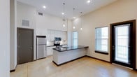 This single-story unit showcases a thoughtfully designed open-concept layout with striking soaring ceilings that lend a sense of spaciousness throughout the living and kitchen areas. Polished concrete floors run continuously underfoot, complementing the unit's contemporary industrial aesthetic.

The kitchen is a standout feature, centered around a large peninsula with dark granite countertops and an undermount sink, illuminated by a trio of sleek pendant lights overhead. White shaker-style cabinetry extends floor to ceiling along the back wall, providing generous storage, while a full suite of stainless steel appliances — including a refrigerator, electric range, and built-in microwave — completes the space. A subway tile backsplash adds a clean, modern finish. Natural light enters through multiple windows fitted with blinds, brightening both the kitchen and living areas.

The entryway features two solid gray doors — one serving as the unit entry and one providing access to an interior closet or storage area. The bathroom includes a full tub and shower combination with a white surround, granite vanity countertop, and recessed lighting. A dedicated laundry area houses a stacked washer and dryer for added convenience.