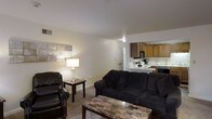 This single-floor space is designed with an open-concept layout that integrates the living area and kitchen into one cohesive space. The living room is carpeted and comfortably sized, providing enough room for a full sofa, a reclining armchair, a side table, and a coffee table with a stone-like surface. Neutral-toned walls keep the space feeling bright and clean, complemented by overhead lighting and a table lamp that add warmth to the room. A decorative wall piece adds a subtle artistic touch above the seating area.

The kitchen is visible from the living room and features warm wood-toned cabinetry in both upper and lower configurations, offering ample storage. It is equipped with standard appliances, including a microwave, stovetop range, and additional countertop appliances, making it a practical space for everyday cooking. The kitchen benefits from its own overhead lighting and light-colored countertops that contrast nicely with the cabinetry. A hallway visible in the background suggests access to additional rooms within the unit. Overall, the space presents a functional and straightforward layout suited for everyday living.