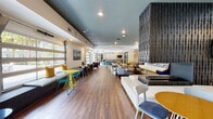 Located at 1903 SW 14th Terrace in Gainesville, Florida, this single-floor community clubhouse is a dynamic, thoughtfully designed gathering space that blends modern aesthetics with functional versatility. The interior is unified by warm wood-look flooring that runs throughout, complemented by a dark painted ceiling that adds contrast and depth to the open layout.

At the entrance, a striking geometric-patterned tile floor welcomes visitors alongside a wood-paneled accent wall and vibrant yellow seating. Nearby, a reception counter with bar-height stools and a wall-mounted television serves as an informational hub, flanked by oversized drum pendant lights that illuminate the area with style.

The main lounge stretches expansively and is anchored by a bold three-dimensional textured wall column, separating distinct seating zones. One area features a built-in banquette with a large-screen TV, surrounded by eclectic mix-and-match chairs and tables in teal, yellow, and gray tones. A long communal dining table with industrial-style pendants overhead provides space for group gatherings or collaborative work.

A standout feature is the row of operable glass garage-style doors along one wall, which open to flood the space with natural light and views of the surrounding landscape. Throughout, bold blue accent walls, sculptural light fixtures, and curated furnishings create an energetic yet welcoming atmosphere ideal for socializing and community engagement.