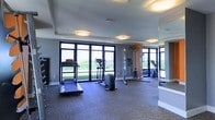 This single-floor fitness space is designed to support a well-rounded workout experience in a clean, functional environment. The room is outfitted with a treadmill, elliptical trainer, and a multi-function cable machine, providing users with both cardio and strength training options. A stationary bike is also visible, reflected in the large wall-mounted mirror that lines one side of the room, effectively expanding the sense of space and allowing users to monitor their form during exercise.

The flooring consists of durable, rubberized gym-grade material in a muted blue-grey tone, which provides cushioning and safety underfoot. Overhead, flush-mounted ceiling lights offer even illumination throughout the space. The most notable architectural feature is the row of large, floor-to-ceiling windows with dark-framed mullions along the exterior wall, flooding the room with natural light and providing open views of a green outdoor landscape. The grey-toned walls complement the overall modern aesthetic, creating a bright, motivating atmosphere well-suited for daily fitness use.