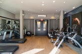 Located at 5 Freer St in Lynbrook, NY, this modern fitness center offers a well-appointed, single-floor workout facility designed with both functionality and aesthetics in mind. The space features light hardwood-style flooring throughout, complemented by a clean, neutral color palette accented by bold teal walls that add a dynamic visual energy to the environment. Recessed lighting and large floor-to-ceiling windows and glass doors flood the interior with abundant natural light, while providing direct views and access to an outdoor courtyard area. The facility is equipped with a comprehensive selection of cardio machines, including treadmills, stationary bikes, rowing machines, and elliptical trainers, all from leading brands such as Life Fitness. A dedicated stretching and functional training area features carpeted flooring, stability balls, and resistance equipment. Full-length mirrors along one wall visually expand the space and assist with form during workouts. Bold, large-scale fitness-themed artwork adorns the teal accent walls, contributing to a motivating atmosphere. A dumbbell rack and additional free weights are also available for strength training. The space is finished with a modern double-door entry, climate control, and a thoughtful open layout that ensures comfortable circulation throughout the facility.