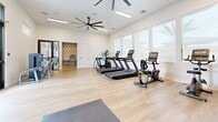 Located in Phoenix, AZ, this fitness center offers a well-designed, single-level exercise space with a thoughtful layout that maximizes functionality. The room features light hardwood-style flooring throughout, crisp white walls, and elevated ceilings equipped with large ceiling fans and recessed lighting panels, contributing to an open, airy atmosphere.

Natural light floods the space through oversized windows along one wall, offering views of palm trees and the surrounding Arizona landscape. Glass double doors provide an additional entry point and further enhance the connection to the outdoors.

The cardio zone is well-stocked with multiple treadmills, an elliptical trainer, and a recumbent stationary bike. A dedicated strength-training area includes a Matrix dual-cable functional trainer with an adjustable bench, a selectorized weight machine, and a rack of dumbbells. Exercise mats on the floor provide space for stretching or floor-based workouts.

A bold red accent wall with framed artwork and a wall-mounted flat-screen television adds a dynamic visual element, while a large mirror expands the sense of depth. A water fountain station is conveniently integrated into the space. The overall design balances a modern, energetic aesthetic with practical fitness amenities.