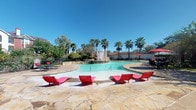 This beautifully designed outdoor amenity space offers a resort-like atmosphere centered around a generously sized swimming pool with a striking central fountain feature. The pool is surrounded by a flagstone deck finished in warm earth tones, furnished with bold red chaise lounges that create a vivid contrast against the sparkling turquoise water. Red market umbrellas provide additional shaded seating areas along the pool's perimeter, making it an ideal setting for relaxation.

Tall, swaying palm trees line the pool area, lending a tropical character to the space and framing unobstructed views of open blue skies. Lush, mature landscaping — including ornamental shrubs, grasses, and large shade trees — borders the walking paths that wind throughout the grounds, creating a sense of privacy and natural beauty.

A standout architectural feature is the brick pergola structure, constructed with classic red brick columns and a slatted wooden overhead canopy fitted with ceiling fans for warm-weather comfort. Beneath it, additional lounge chairs offer a shaded alternative for outdoor relaxation. The surrounding grounds are enclosed by wrought-iron fencing, maintaining both security and an open, airy aesthetic. The overall ambiance is inviting, well-maintained, and designed for leisure.
