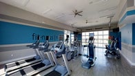 This fitness center offers a well-appointed single-level layout designed to support a comprehensive cardio workout experience. The space is defined by a striking teal accent wall adorned with the word "fitness" in bold lettering, lending the room an energetic and motivating atmosphere. The flooring is light-toned hardwood-style material, contributing to a clean, contemporary aesthetic throughout.

A lineup of high-quality cardio machines — including treadmills, ellipticals, and stair climbers — is arranged in an organized fashion, each equipped with individual digital display screens for tracking workouts. The equipment is positioned to take full advantage of the large garage-style windows along one wall, providing open views to the outdoors and filling the interior with ample natural light.

The ceiling features multiple ceiling fans that aid in air circulation, enhancing comfort during workouts. Recessed and strip lighting supplements the natural light, ensuring the space remains bright at all hours. The overall ambiance is modern, clean, and purpose-driven, making this fitness center a functional and inviting environment for regular exercise.