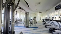 This fitness center is a spacious, single-floor facility designed to support a comprehensive workout experience. The layout is open and airy, with large floor-to-ceiling windows lining the perimeter that flood the space with abundant natural light and offer expansive outdoor views. White walls and a high, curved ceiling contribute to the bright, clean aesthetic, while multiple ceiling fans are evenly distributed throughout to promote comfortable air circulation during workouts. The flooring features a contemporary blue and white patterned carpet, adding a polished, modern touch to the overall design. The facility is outfitted with a diverse selection of professional-grade equipment, including cable and pulley machines, a multi-function weight rack with dumbbells, and a variety of cardio machines such as rowing machines and ellipticals. The equipment is organized across the space to allow for easy navigation and unobstructed movement between stations. Mirrors are strategically placed to enhance the sense of space and support proper form during exercise. The overall ambiance is clean, modern, and welcoming, making this fitness center a well-appointed amenity suitable for a wide range of fitness levels and training styles.