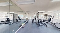 The Pelican Watch Fitness Center in Virginia Beach, Virginia, is a bright and functional single-floor facility designed to support a well-rounded fitness routine. The space is finished with durable, cushioned blue-gray carpet flooring throughout, white walls, and a textured ceiling with flush fluorescent lighting that keeps the environment clean and well-lit.

The facility is generously equipped with a diverse range of exercise equipment, including treadmills, elliptical trainers, upright and stationary bikes, a multi-station cable weight machine, a weight bench with free weights, a rowing attachment, and an assortment of dumbbells and stability balls. A wall-mounted television provides entertainment during workouts.

Full-length mirrors line one wall, visually expanding the space and allowing users to monitor their form during exercise. Multiple large windows wrap around the room on two sides, flooding the interior with natural light and offering pleasant views of both the outdoor pool area — with its vibrant teal umbrellas — and the surrounding community grounds. A glass-paned door provides direct access to the exterior.

The overall ambiance is clean, open, and welcoming, making the Pelican Watch Fitness Center a practical and comfortable destination for residents seeking a convenient on-site workout experience.