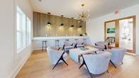 This thoughtfully designed single-floor communal space offers an open-concept layout that seamlessly integrates a living room, dining area, and kitchen into one cohesive environment. Light wide-plank hardwood flooring flows throughout, lending warmth and continuity across the entire space.

The living area is a standout, anchored by a dramatic slatted wood accent wall that serves as a striking backdrop to a built-in counter with bar seating and pendant lighting. A statement sputnik chandelier hangs overhead, illuminating a curated seating arrangement of mid-century modern upholstered chairs surrounding a minimalist coffee table.

The dining zone features tall bistro-style tables and bar stools, positioned near glass double doors that open to an outdoor area, allowing abundant natural light to fill the room through multiple windows.

The kitchen is well-appointed with rich, warm-toned shaker-style cabinetry, white quartz countertops, stainless steel appliances including a range, microwave, and refrigerator, and a tile backsplash. A central kitchen island with seating provides additional prep and gathering space. A set of wood-framed glass doors leads to an adjacent private room, adding functional flexibility to the layout. The overall ambiance is clean, modern, and inviting.