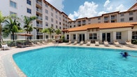 This inviting outdoor amenity space showcases a generously sized swimming pool as its centerpiece, with crystal-clear blue water that evokes a true resort atmosphere. The pool deck is finished with light-toned pavers and lined with a row of cushioned lounge chairs, offering ample space for relaxation and sunbathing. A charming poolside pavilion or cabana structure with a distinctive terra cotta tile roof provides shade and a comfortable retreat, complementing the Mediterranean-inspired architectural style of the surrounding multi-story building. The building itself features light stucco exteriors, private balconies, and large windows that overlook the pool area. Tall palm trees and tropical greenery frame the space, reinforcing a warm, sun-drenched ambiance. A secure fencing perimeter encloses the pool area, providing both safety and privacy. The open-sky setting allows for abundant natural light throughout the day, making this a bright and welcoming communal outdoor space ideal for leisure and socializing. The overall design blends practical amenities with an aesthetic that reflects a relaxed, tropical lifestyle.