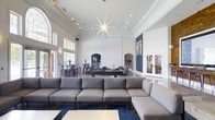 This single-floor communal space is designed for socializing and recreation, offering an open, airy layout with impressively high ceilings that amplify the sense of spaciousness throughout. The main lounge area is anchored by a large, plush sectional sofa in neutral gray tones, set atop a deep navy blue area rug that grounds the space with a bold pop of color. Above, a striking starburst chandelier serves as a dramatic focal point, casting a warm, inviting glow across the room.

Beyond the seating area, a full-size pool table takes center stage, making it an ideal gathering spot for friendly competition. The space transitions seamlessly into a bar and dining area, where bar-height tables paired with vibrant purple chairs add a lively, modern flair. Arched passageways lend architectural charm and visual interest, while a decorative textured panel serves as an elegant room divider.

Natural light pours in through tall windows and glass-paneled doors, connecting the interior to the outdoors and enhancing the bright, welcoming atmosphere. Curated artwork adorns the walls alongside thoughtful accent lighting, contributing to a polished yet comfortable ambiance. Overall, the space is well-suited for both casual relaxation and lively social gatherings.