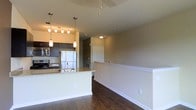This single-floor space showcases a well-designed open-concept layout that seamlessly connects the kitchen and living areas. The kitchen is equipped with stainless steel appliances, including a full-size refrigerator and stove, complemented by dark cabinetry that provides a sleek, modern contrast against the warm-toned walls. A peninsula with a granite countertop serves as both a functional workspace and a casual dining or gathering area, with a built-in sink adding to its practicality. Pendant lighting hangs above the peninsula, while a track lighting fixture illuminates the kitchen, ensuring the space is well-lit throughout. The living area features rich, dark hardwood flooring that extends throughout, adding warmth and depth to the interior. A half-wall with white trim defines the boundary of the living space, contributing a clean architectural detail. Neutral beige walls provide a versatile backdrop, while flush-mount ceiling lighting ensures consistent illumination. A doorway visible toward the rear of the space leads to additional rooms, suggesting a functional and thoughtfully arranged floor plan overall.
