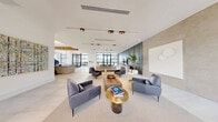 Located at 8500 N Sherman Circle in Miramar, FL, this sophisticated clubhouse offers a refined and welcoming environment designed for community gathering and relaxation. The space is spread across a single level and presents an open, flowing layout that connects its distinct functional areas seamlessly.

The grand entrance foyer immediately sets the tone with soaring double-height ceilings, expansive floor-to-ceiling black-framed glass doors, and large-format light stone tile flooring that carries throughout the space. Abundant natural light pours in through the oversized windows, offering views of lush Florida landscaping including swaying palms.

The main lounge area is elegantly appointed with contemporary furniture, including leather accent chairs and plush sofas arranged around a rustic wood coffee table. A sleek, dark-paneled feature wall houses a flat-screen TV, while track lighting above adds a warm, curated ambiance. Tall indoor trees and sculptural décor bring organic warmth to the modern aesthetic.

A dedicated dining and social area showcases exposed reclaimed wood ceiling beams set against white tongue-and-groove ceilings with cove lighting, creating a striking architectural contrast. A long communal dining table is complemented by a built-in catering counter and open shelving, making it ideal for events and gatherings. Sheer drapery softens the natural light in this inviting area, completing the space's polished yet comfortable character.