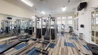 Located at 30685 FM2978 in Magnolia, Texas, this single-story community facility is designed with both comfort and functionality in mind. The space showcases light hardwood flooring throughout, crisp white walls, and soaring ceilings that create an open, airy atmosphere.

The central lounge area serves as the heart of the space, featuring a tufted charcoal sofa, ceiling fan, recessed lighting, and a wall-mounted flat-screen TV above a built-in gas fireplace. An abundance of multi-pane windows floods the room with natural light and provides views of the surrounding landscape. Elegant table lamps and curated artwork add warmth to the contemporary décor.

The kitchen is a standout feature, highlighted by a large granite island with bar seating, white shaker cabinetry, a classic subway tile backsplash, stainless steel appliances, and striking geometric pendant lights overhead. A dedicated coffee and beverage station is conveniently positioned nearby.

A private office or workroom offers built-in shelving with arched detailing, a granite-topped wall desk, and a pendant light, creating a productive and polished workspace. An additional room with glass-paned French doors appears to function as a fitness or multipurpose area. Arched entryways connect the various zones, lending architectural character throughout this thoughtfully designed community space.