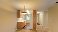This single-floor space offers a functional and cohesive layout, with the kitchen serving as a central highlight. The kitchen is outfitted with light honey-toned wood cabinetry that provides ample storage, complemented by clean white countertops and a built-in sink positioned beneath a window that allows natural light to filter in. A standout pendant light fixture hangs overhead, adding a warm, inviting focal point to the space. A distinctive wood-wrapped structural column adds architectural character, visually anchoring the transition between the kitchen and the adjoining areas. The flooring transitions between warm hardwood tones throughout, contributing to a sense of continuity across the space. Recessed and flush-mount ceiling lighting ensures consistent illumination throughout. A hallway extending from the main area leads to additional rooms, each accessible through white paneled doors. The overall color palette — soft beige walls, natural wood tones, and white trim — creates a clean, neutral ambiance that is both practical and welcoming.