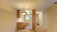This single-floor space offers a functional and cohesive layout, with the kitchen serving as a central highlight. The kitchen is outfitted with light honey-toned wood cabinetry that provides ample storage, complemented by clean white countertops and a built-in sink positioned beneath a window that allows natural light to filter in. A standout pendant light fixture hangs overhead, adding a warm, inviting focal point to the space. A distinctive wood-wrapped structural column adds architectural character, visually anchoring the transition between the kitchen and the adjoining areas. The flooring transitions between warm hardwood tones throughout, contributing to a sense of continuity across the space. Recessed and flush-mount ceiling lighting ensures consistent illumination throughout. A hallway extending from the main area leads to additional rooms, each accessible through white paneled doors. The overall color palette — soft beige walls, natural wood tones, and white trim — creates a clean, neutral ambiance that is both practical and welcoming.