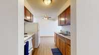 This single-floor, 2-bedroom, 1-bathroom unit offers a straightforward and functional layout designed for efficient living. The galley-style kitchen is lined with warm wood cabinetry on both sides, providing ample upper and lower storage, and is equipped with a double-basin sink, electric range, and a full-size refrigerator. Light wood-look flooring runs throughout the kitchen, which opens toward the living area, creating a connected feel. A ceiling fan with a light fixture adds airflow and illumination to the space.

The living room is a generously sized, open area with light carpet, neutral walls, and a wall-mounted air conditioning unit, offering a comfortable retreat. Both bedrooms are carpeted and feature baseboard heating and windows with horizontal blinds that allow natural light while maintaining privacy. The primary bedroom includes a closet with built-in shelving, while the second bedroom also features a closet and clean, neutral finishes throughout.

The bathroom is simply appointed with a full tiled tub-and-shower surround in classic white, complemented by matching white fixtures. Hallways connecting the rooms provide a logical flow through the unit. Overall, the space presents a clean, no-frills interior with practical amenities suited for comfortable, day-to-day living.